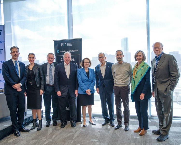 Nine academics pose standing side by side while smiling for the camera in a room with a glass wall and the Boston skyline behind them