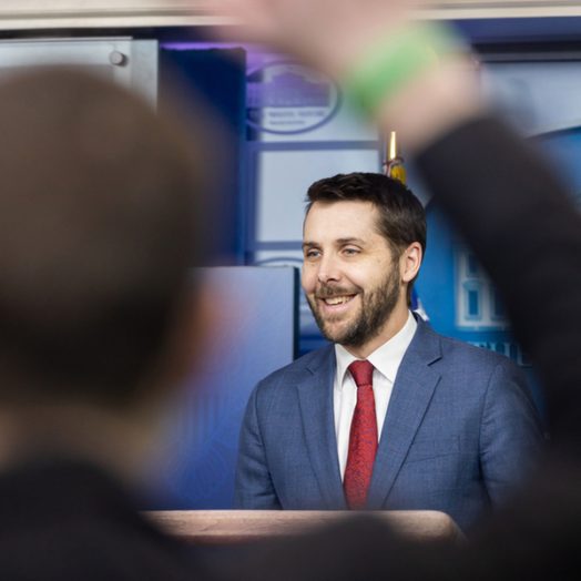 Brian Deese speaks in the White House Briefing Room. In the blurry foreground, a person raises their hand.