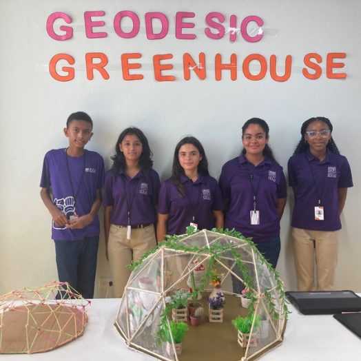 Five children wearing purple shirts stand against a wall displaying the words “Geodesic Greenhouse.” Two geodesic models are on a table in front of them.
