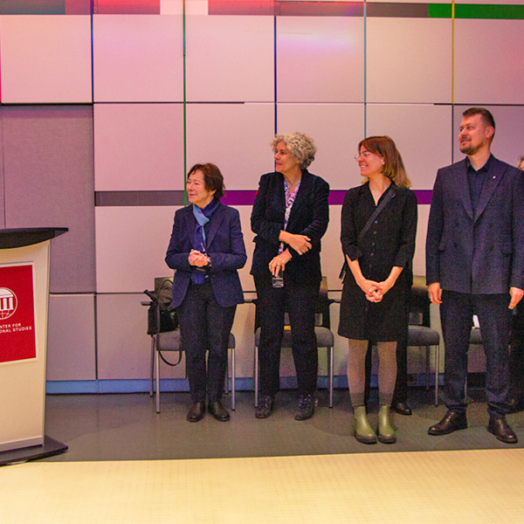 At left, Cynthia Barnhart speaks at a podium while about half a dozen indiviuals look on. The event is in a room lined with white square tiles and colored accent tiles.