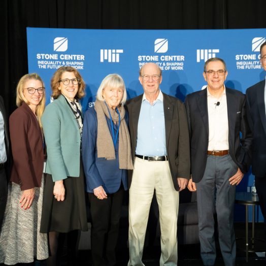 Seven people stand smiling in front of a blue MIT Stone Center backdrop