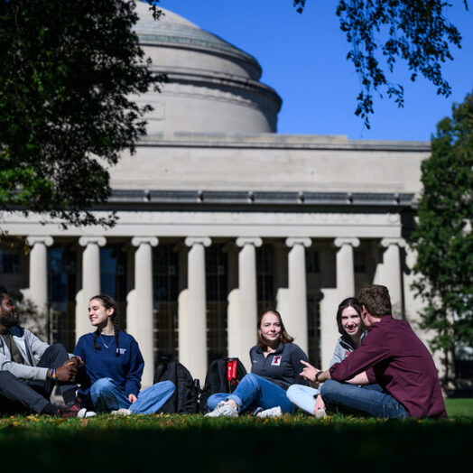 Four students sit on a grass courtyard on a sunny, early fall day. The MIT dome is in the background.