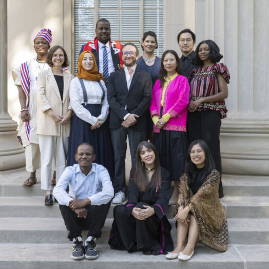 12 people, some with traditional dress from various countries, pose on the steps of an MIT building with columns behind them