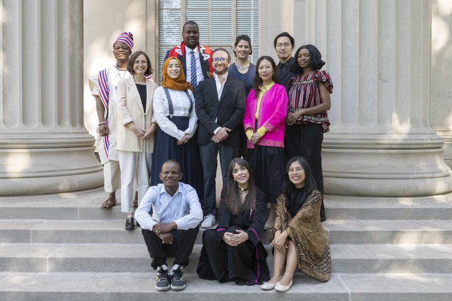 12 people, some with traditional dress from various countries, pose on the steps of an MIT building with columns behind them