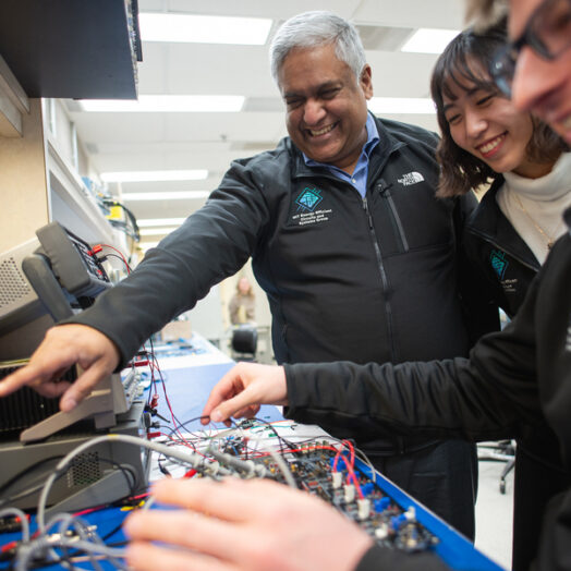 Anantha Chandrakasan smiles and points to a computer chip, alongside two grinning graduate students