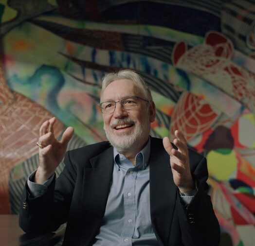 Duane Boning, seated in front of a colorful mural, gestures with both hands