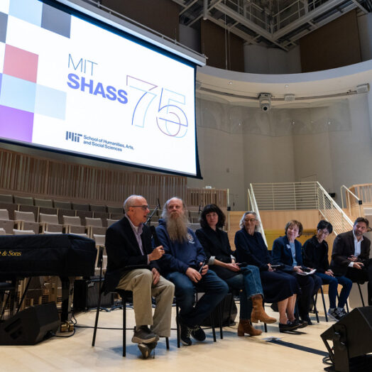 In an auditorium, 7 professors sit in a line beneath a large screen that says SHASS 75